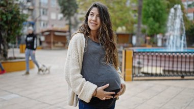 Genç hamile kadın gülümsüyor. Parkta göbeğine dokunuyor.