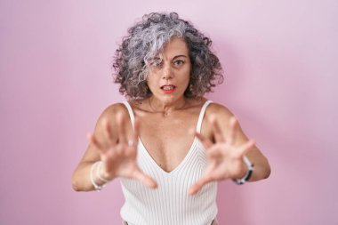 Middle age woman with grey hair standing over pink background afraid and terrified with fear expression stop gesture with hands, shouting in shock. panic concept. 