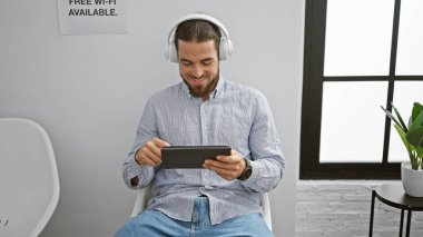 Dokunmatik ped ve kulaklık kullanan genç İspanyol adam bekleme odasında oturmuş gülümsüyor.