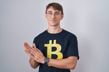 Beyaz, sarışın, bitcoin tişörtlü, mutlu ve neşeli elleri alkışlayan, gururla gülümseyen bir adam. 