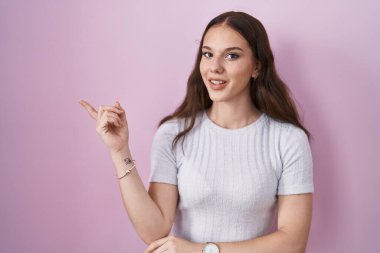 Pembe arka planda duran genç İspanyol kız yüzünde büyük bir gülümsemeyle, kameraya el ile işaret ederek bakıyor.. 