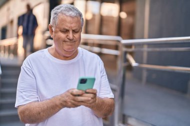 Orta yaşlı, kır saçlı, kendine güvenen, caddede akıllı telefon kullanan bir adam.