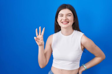 Young caucasian woman standing over blue background showing and pointing up with fingers number three while smiling confident and happy. 