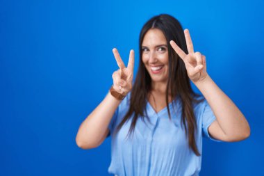 Mavi arka planda dikilen esmer kadın iki elini de zafer işareti yaparak parmaklarını göstererek gülümsüyor. İki numara.. 