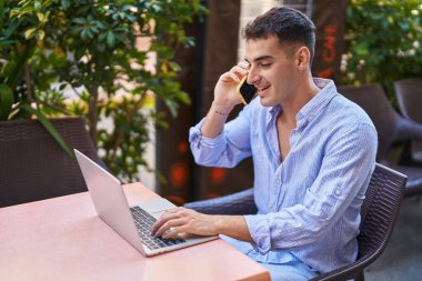 Young hispanic man using laptop talking on smartphone sitting on table at coffee shop terrace