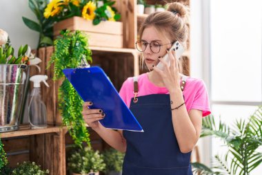 Çiçekçide akıllı telefondan konuşan sarışın kadın pano okuyor.