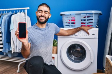 Orta Doğulu sakallı bir adam akıllı telefon ekranı ve çamaşır makinesi gösteriyor. Kameraya seksi bir ifadeyle bakıyor, neşeli ve mutlu bir yüzle.. 