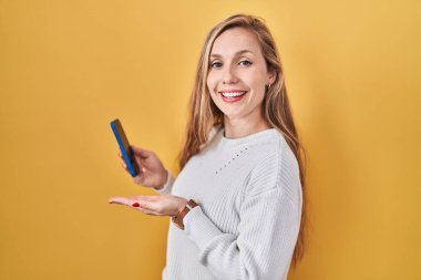 Akıllı telefon kullanarak mesaj yazan genç sarışın kadın avuç içi açık bir şekilde fotokopi alanını gösteriyor, gülümseyerek mutlu bir gülümseme veriyor. 