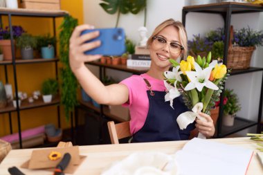 Genç sarışın kadın çiçekçi çiçekçide elinde çiçeklerle akıllı telefonuyla selfie çekiyor.