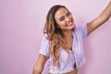 Young hispanic woman standing over pink background dancing happy and cheerful, smiling moving casual and confident listening to music 