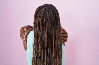 Afro-Amerikalı bir kadın pembe arka planda dikilip kendine sarılıyor, mutlu ve pozitif bir şekilde. Sevgi ve özgüven. 