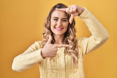 Sarı arka planda duran genç beyaz kadın gülümsüyor ve mutlu yüzlü eller ve parmaklarla çerçeve yapıyor. yaratıcılık ve fotoğrafçılık kavramı. 