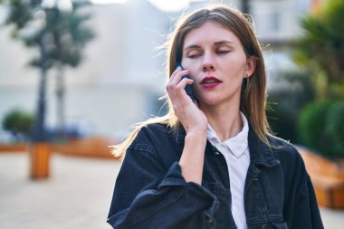 Young blonde woman talking on smartphone with serious expression at park