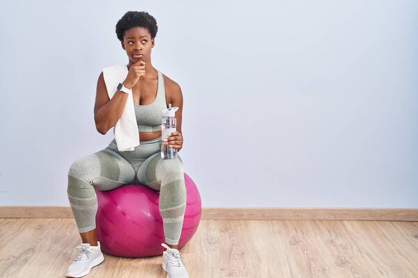 African american woman wearing sportswear sitting on pilates ball with hand on chin thinking about question, pensive expression. smiling with thoughtful face. doubt concept. 