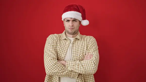 若いですヒスパニック男立って動揺身に着けているクリスマス帽子上の隔離された赤の背景 — ストック写真
