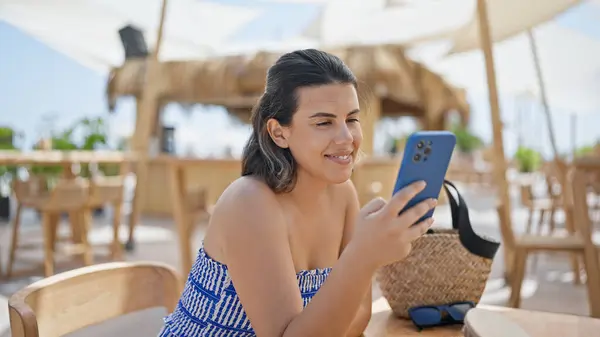 Young hispanic woman smiling happy using smartphone sitting on the table at sunny restaurant terrace