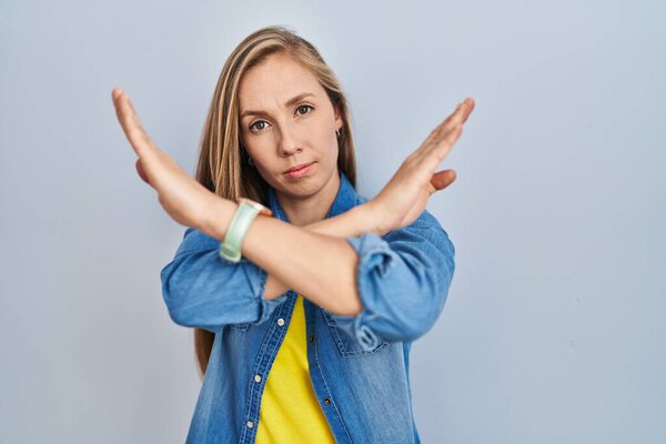 Young blonde woman standing over blue background rejection expression crossing arms doing negative sign, angry face 