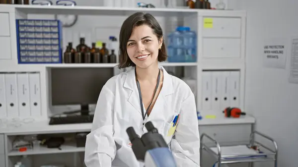 Smiling young hispanic woman scientist joyfully working with microscope in the lab, exemplifying confident, beautiful science research