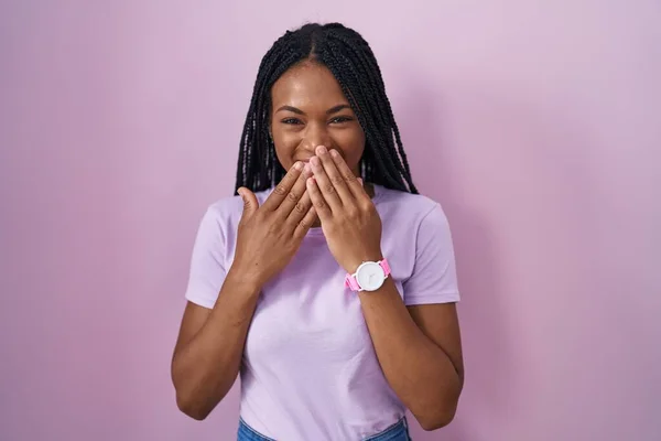 African american woman with braids standing over pink background laughing and embarrassed giggle covering mouth with hands, gossip and scandal concept