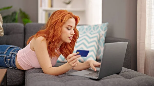Young redhead woman shopping with laptop and credit card lying on sofa at home