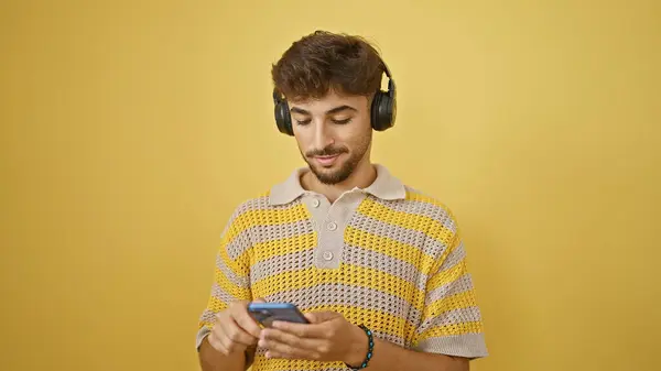 Cool young arab man, with sleek beard, seriously immersed in texting and tuning into music, using the marvel of technology, a smartphone, casually standing against a vivid isolated yellow background