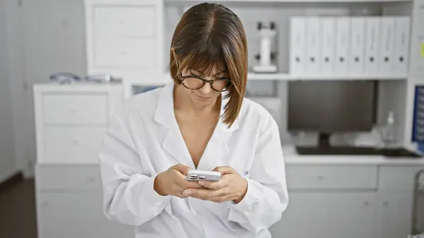 In the hub of science, a young and serious hispanic female scientist, with short hair and a professional demeanor, immersed in beautiful work, tapping away on her smartphone in the lab.