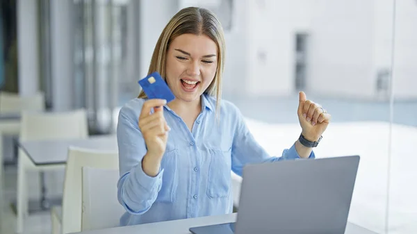 Young blonde woman business worker using laptop and credit card with winner gesture at office