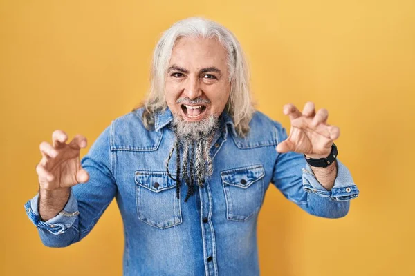 Middle age man with grey hair standing over yellow background smiling funny doing claw gesture as cat, aggressive and sexy expression