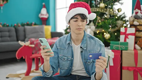 自宅でクリスマスを祝うスマートフォンとクレジットカードで買い物をする若いヒスパニックの男 — ストック写真