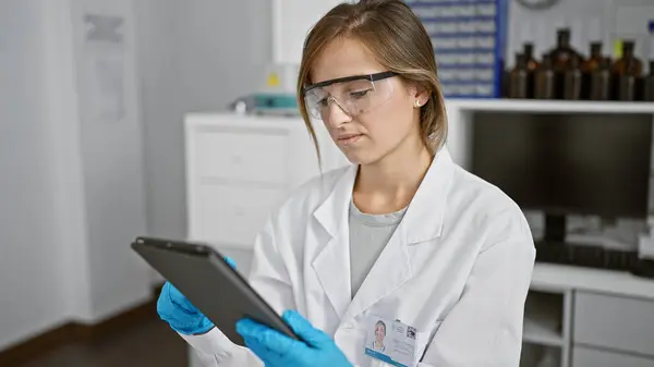 Intrigued young blonde scientist, engrossed in medical analysis, shows off high-tech touchpad in lab bustling with scientific research