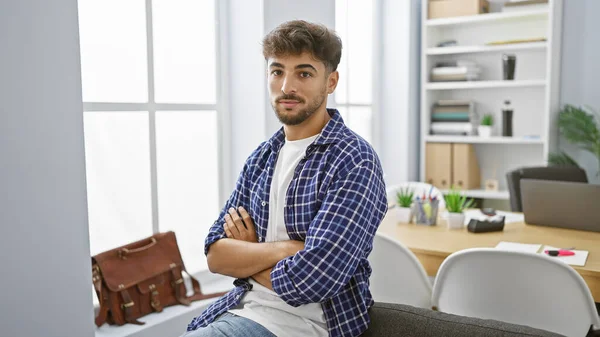 Confident young arab man exhibits professional success, positively embracing his job as he sits in the office with crossed arms gesture