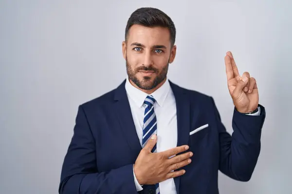 Handsome hispanic man wearing suit and tie smiling swearing with hand on chest and fingers up, making a loyalty promise oath