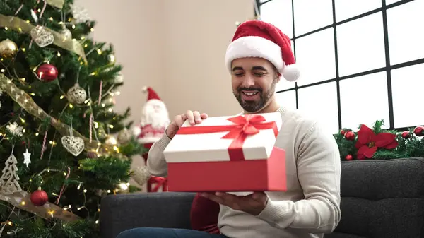 Νεαρός Ισπανός Ξεπακετάρει Δώρο Καθισμένος Στον Καναπέ Δίπλα Στο Χριστουγεννιάτικο — Φωτογραφία Αρχείου