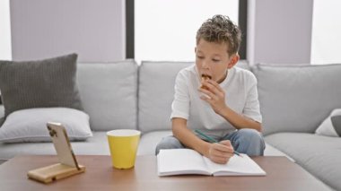 Sevimli sarışın çocuk telefonda ciddi ciddi notlar alıyor, kanepede kapalı otururken tatlı kahvaltıya dalmış, rahat bir evde çevrimiçi dünyaya dalmış.