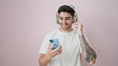 Genç İspanyol adam akıllı telefon ve kulaklık kullanıyor müzik dinliyor ve pembe arka planda dans ediyor.