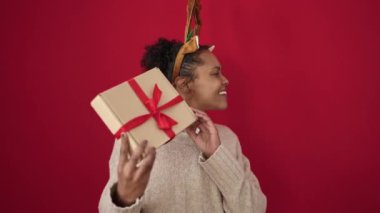 Afrika kökenli Amerikalı bir kadın Noel hediyesi dinliyor. Geyik kulakları takıyor ve izole edilmiş kırmızı arka planda.