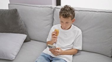 Sevimli sarışın çocuk, akıllı telefonuyla video seyretmeye dalmış, tatlı kruvasan atıştırmalıkları yiyor, rahat evinin oturma odasındaki kanepede oturuyor..