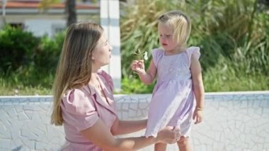 Neşeli beyaz anne ve küçük kızı bir bankta birlikte oturuyor, elinde bir çiçek, neşe ve güven saçıyor.