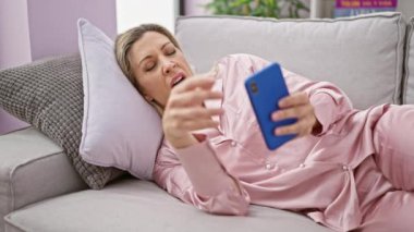 Genç sarışın kadın kanepede uzanıyor ve evde uyumak için akıllı telefon kullanıyor.