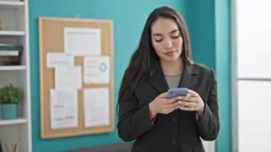 Genç, güzel İspanyol iş kadını. Ofiste akıllı telefon kullanarak gülümsüyor.