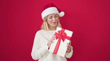 Sarışın, genç bir kadın, Noel şapkası takıyor ve kırmızı arka planda hediyeler tutuyor.