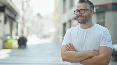 Gri saçlı adam gülümsüyor. Kollarını kavuşturup caddede duruyor.