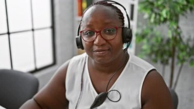 Gülümseyen Afrikalı Amerikalı kadın patron ofiste eğleniyor, kredi kartıyla finans yönetiyor, müşteri hizmetleri desteği için kulaklık takıyor.