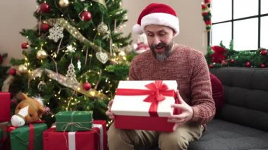 Genç kel adam Noel şapkası takıyor hediyesini açıyor ve evde üzgün görünüyor.
