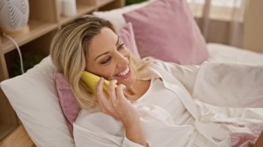 Yatak odasında uzanmış akıllı telefondan konuşan genç sarışın kadın.