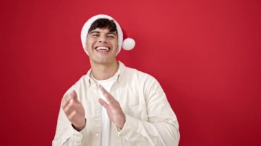 Genç İspanyol adam noel şapkası takıyor, el çırpıyor, kırmızı arka planda alkış tutuyor.