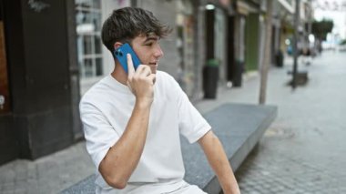 Gündüz Latin kökenli genç bir erkek, güneşli bir şehir sokağında ciddi bir telefon konuşmasına dalmış, havalı bir ifadeyle genç bir adamın hayat tarzı portresi, açık havada akıllı telefonuyla tutkuyla konuşuyor.