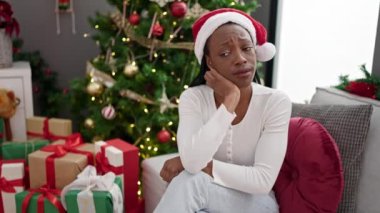 Afrikalı Amerikalı bir kadın Noel ağacının yanında koltukta oturmuş üzgün üzgün bakıyor.