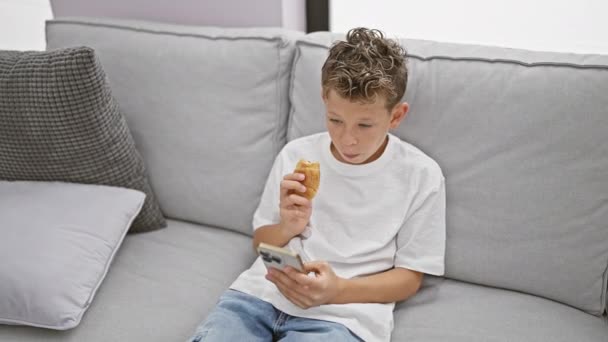 愛らしいブロンドの少年は 彼の居心地の良い家のリビングルームにソファーに座って スマートフォンでビデオを見ることに真剣に興奮しました — ストック動画