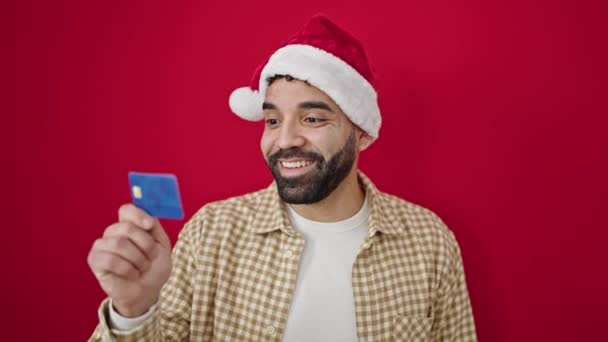 头戴圣诞帽头戴圣诞礼帽的年轻人带着信用卡 背景是孤立的红色 — 图库视频影像